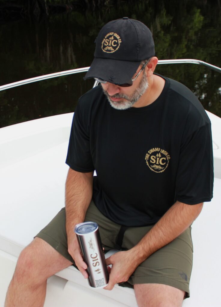 Man wearing black SIC Distressed Ball Cap and black Swamp Iron Co. T-Shirt while seated on a boat